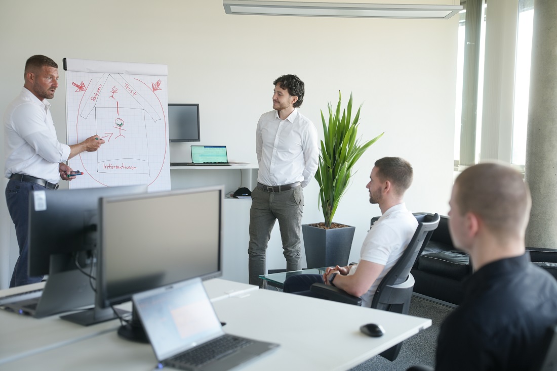 Ein Mann steht vor einem Flipchart und erklärt drei jungen Männern ein Kundenthema.