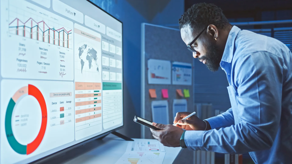 Mitarbeitender steht vor einem Monitor mit Analysen / Grafiken und macht sich Notizen.