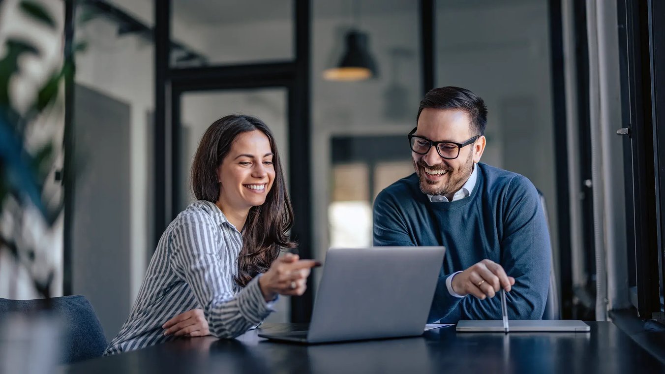 Two colleagues, husband and wife, are sitting at a table looking at a laptop.