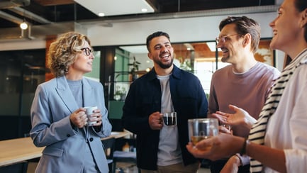 Vier collega's van verschillende generaties staan bij elkaar met een kop koffie en kletsen terloops.