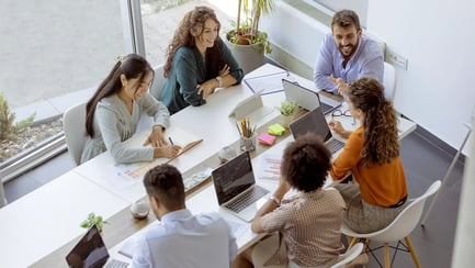Zes jonge studenten zitten aan een groot bureau met een laptop en werken aan een workshoponderwerp.