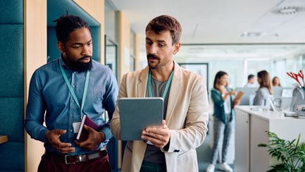 Two businessmen discuss something on an iPad as they walk through the office.