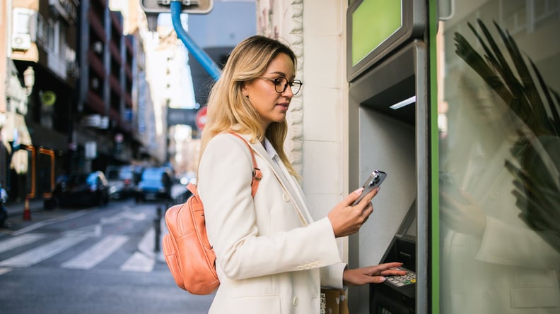 Eine Frau hebt draußen Geld am Bankautomaten ab und schaut dabei auf ihr Handy