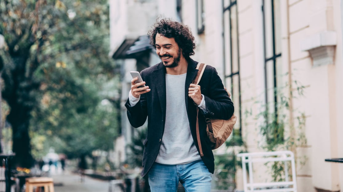 Mann unterwegs im Freien, der auf sein Handy blickt und dabei lächelt.