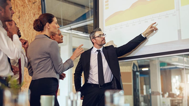 Man in a suit is presenting a chart displayed on a digital screen to a group of attentive colleagues.
