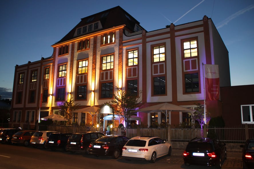 Nighttime view of a two-story European-style building with illuminated windows, a balcony, flag, and parked cars.